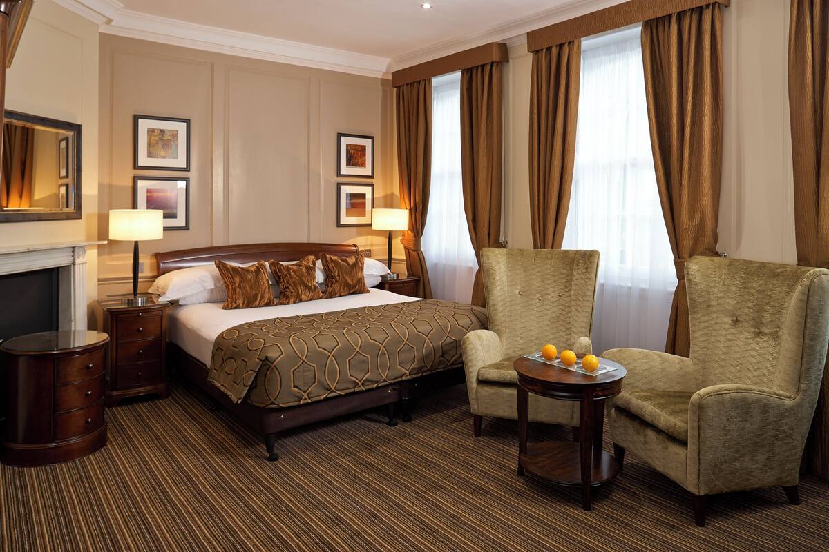 A hotel room with a large bed, two side tables, a chair, a small table with fruit, and a fireplace.