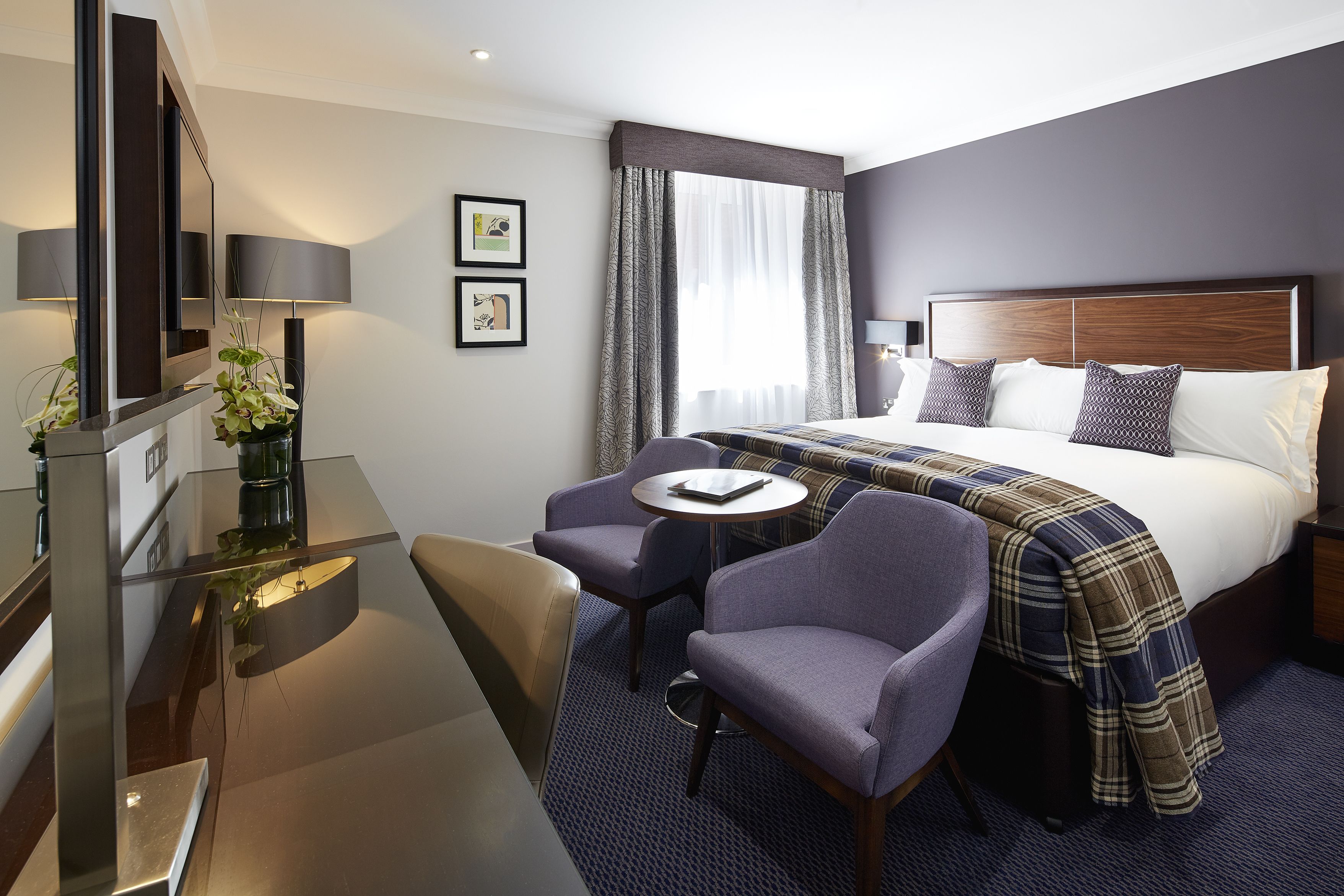 A hotel room with a bed, a desk with a chair, a small table, and a television.
