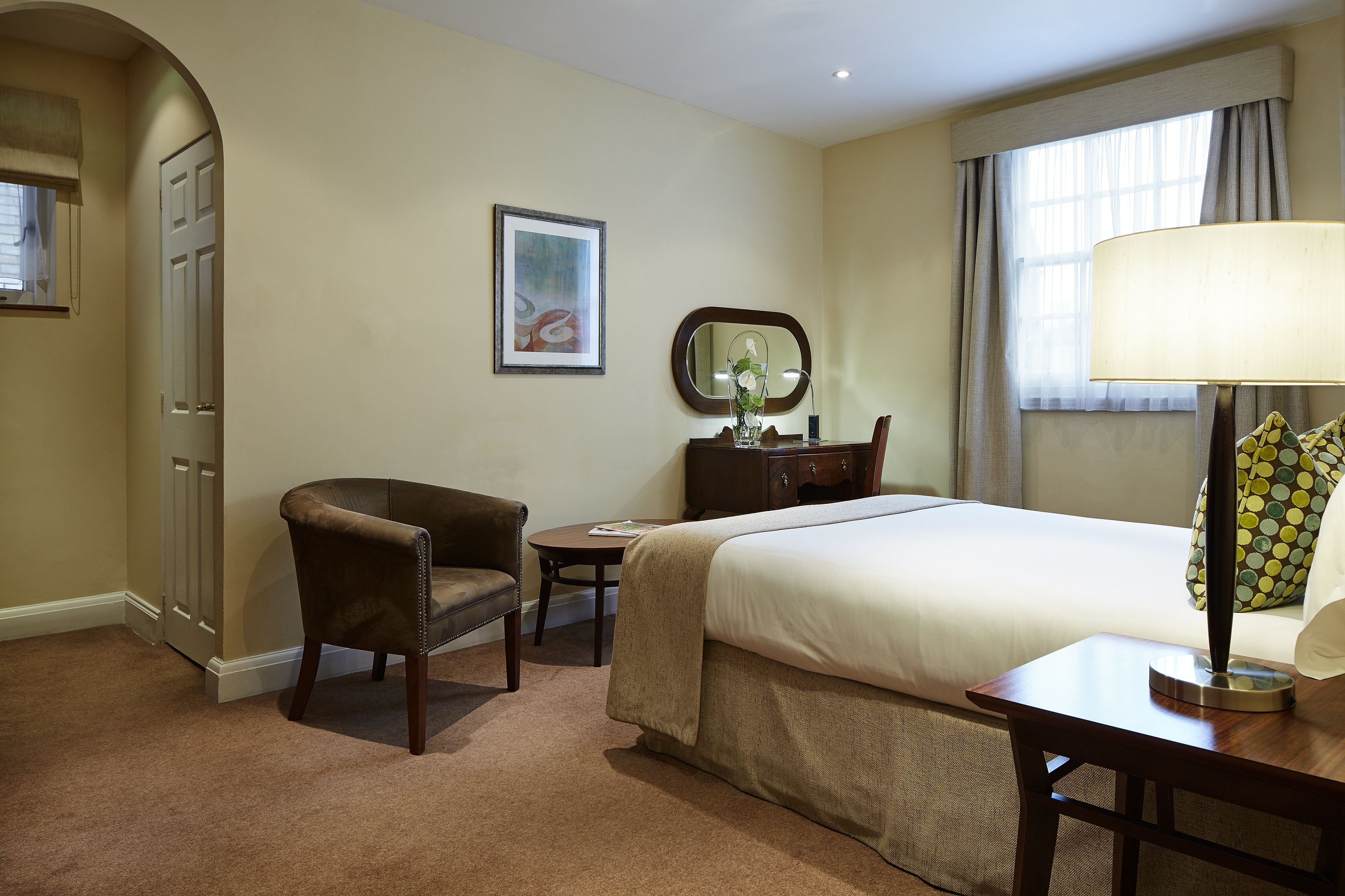 A hotel room with a bed, a chair, a desk, a mirror, and a window with curtains.
