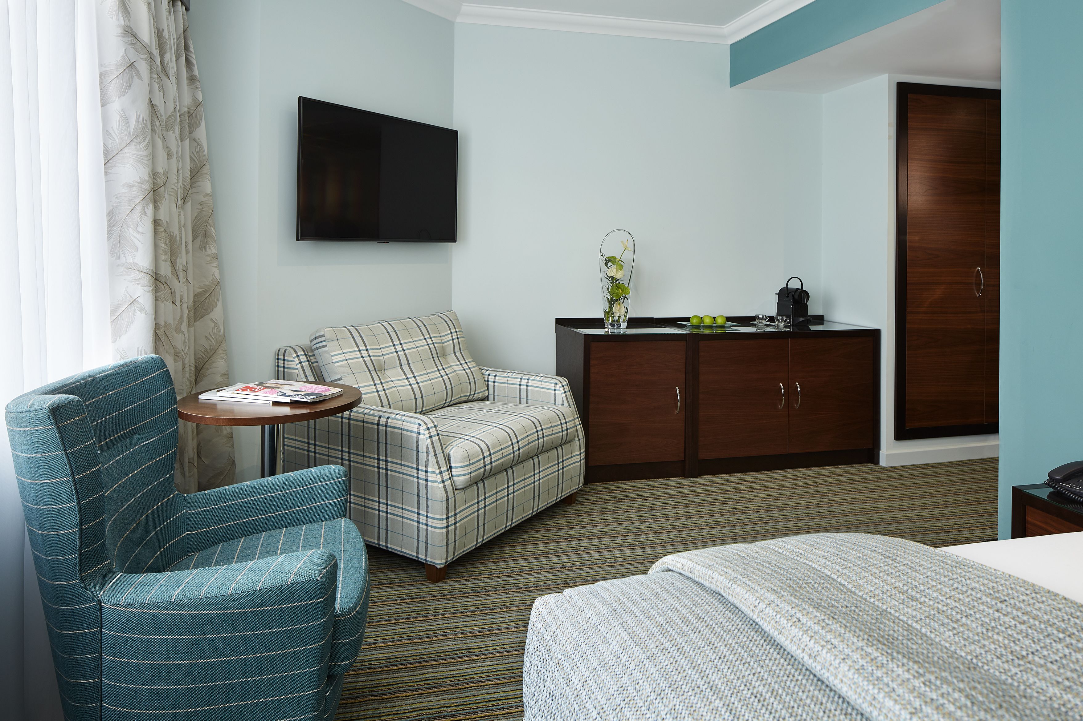 A hotel room with a blue-striped armchair, a plaid sofa, a small round table with a magazine, a TV mounted on the wall, a wooden cabinet, and a bed with a patterned bedspread.