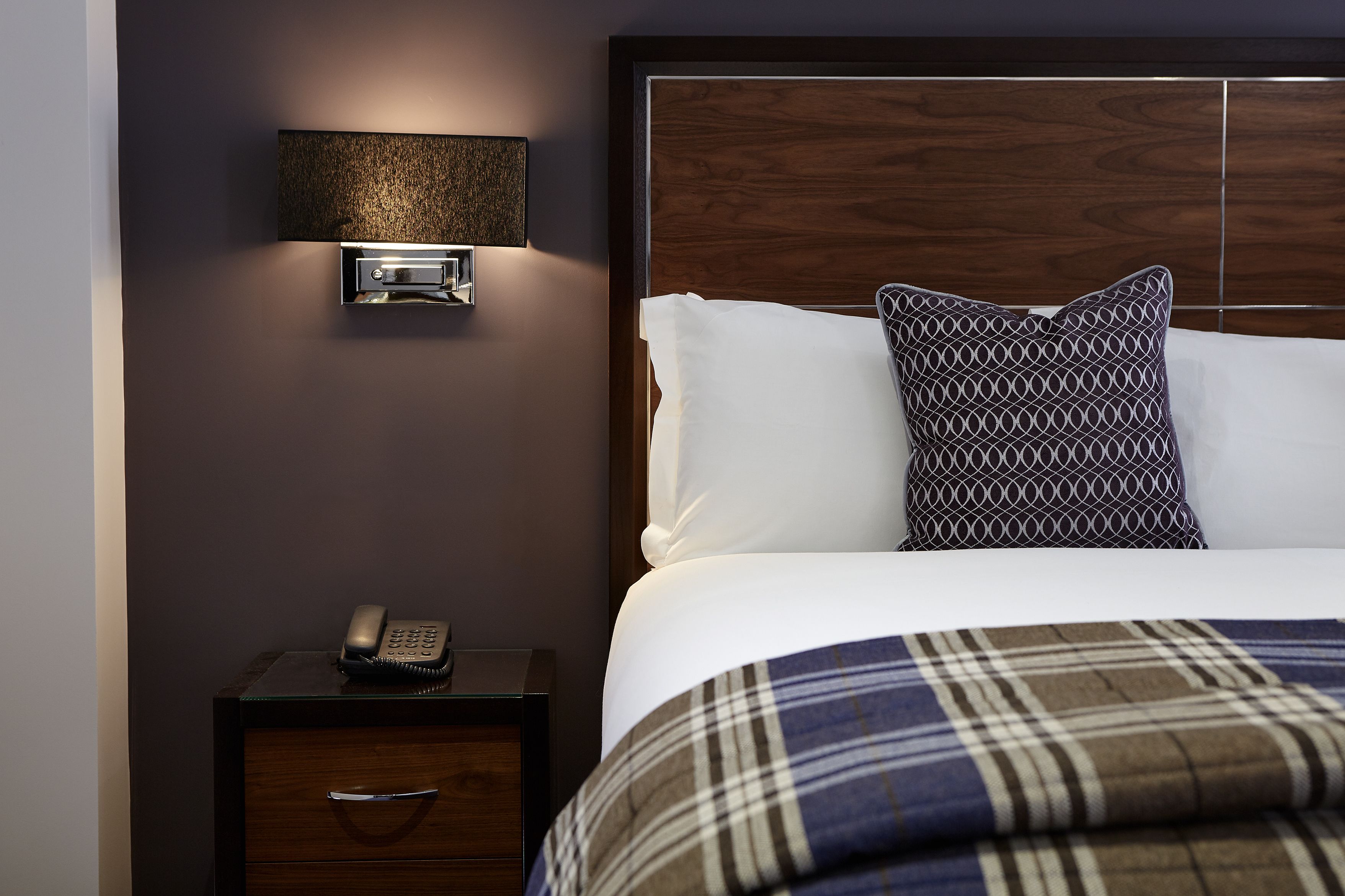 Habitación de hotel con cabecera de madera, mesita de noche con teléfono y manta de cuadros sobre la cama.