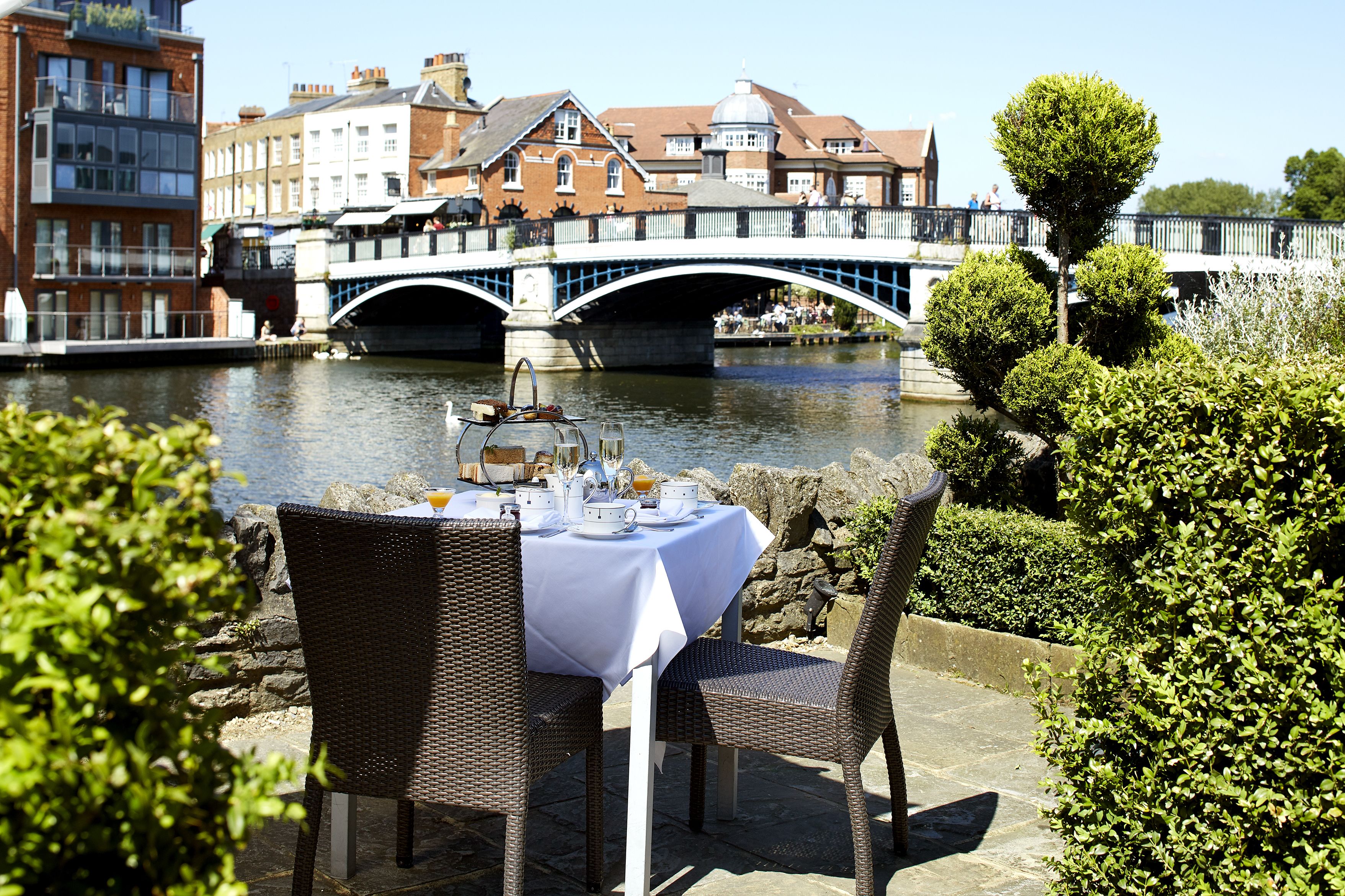 An outdoor dining area with a table set for two, overlooking a river with a bridge in the background.
