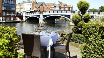 An outdoor dining area with a table set for two, overlooking a river with a bridge in the background.