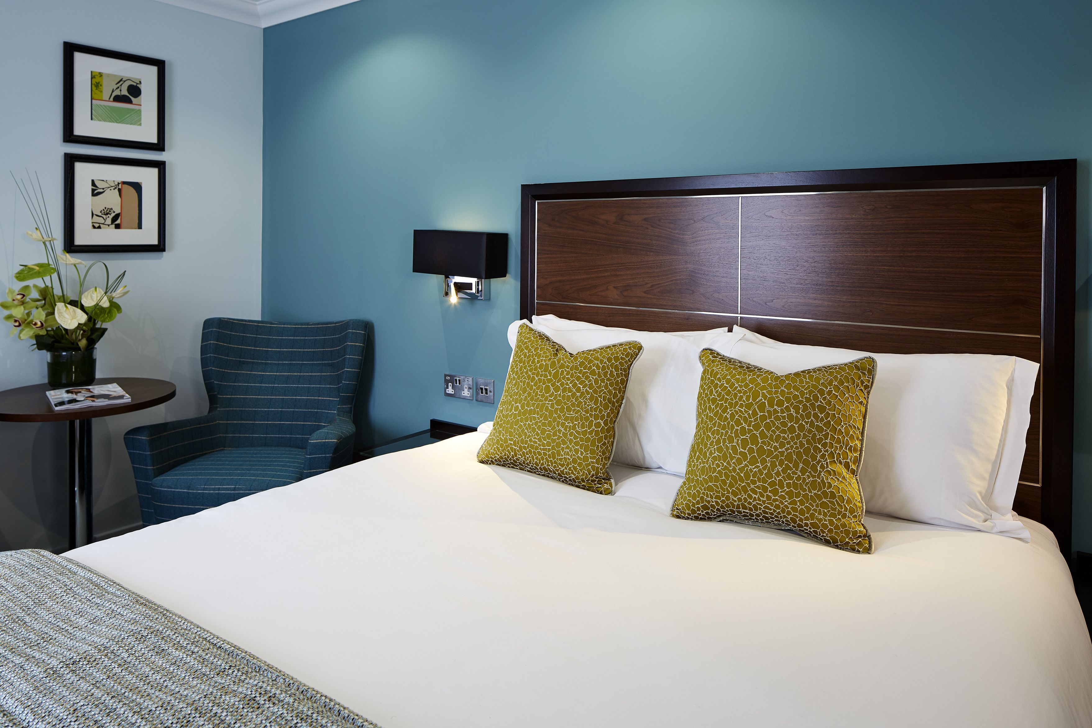 Ein Hotelzimmer mit einem großen Bett, zwei grün gemusterten Kissen, einem blauen Sessel, einem kleinen Tisch mit einer Zeitschrift und zwei gerahmten Bildern an der Wand.