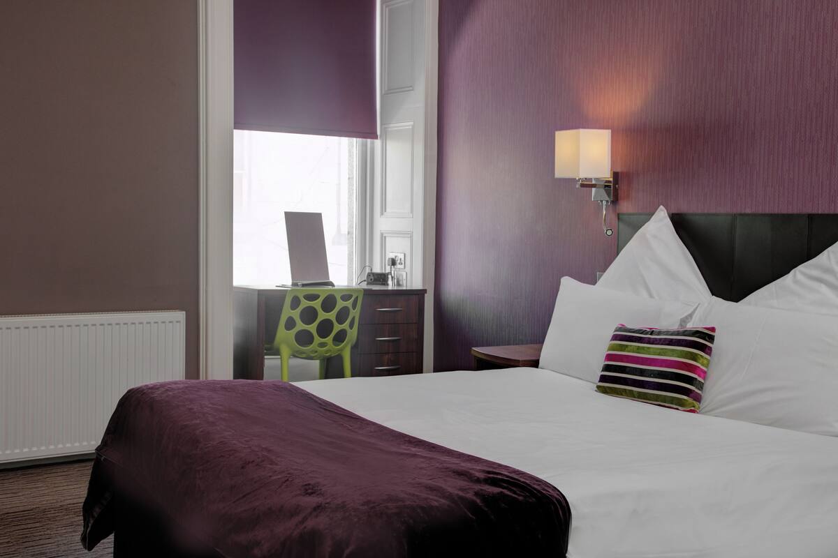 A hotel room with a bed, a desk, a chair, and a lamp.