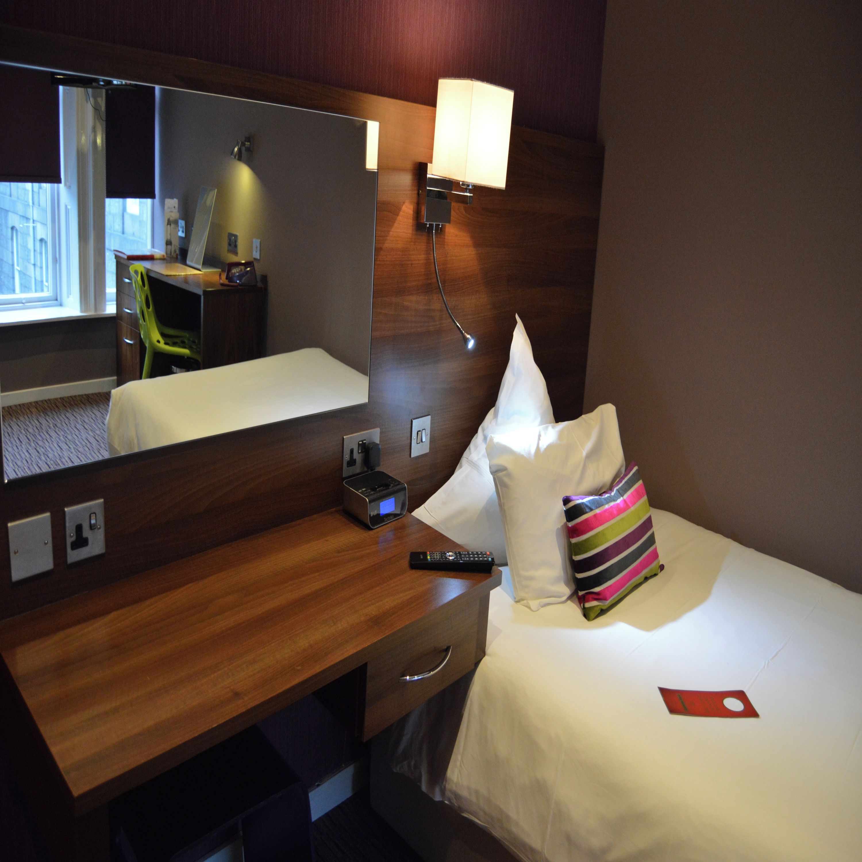 Een hotelkamer met een houten bureau, een spiegel, een bedlampje, een bed met witte lakens, een kleurrijk gestreept kussen en een afstandsbediening.