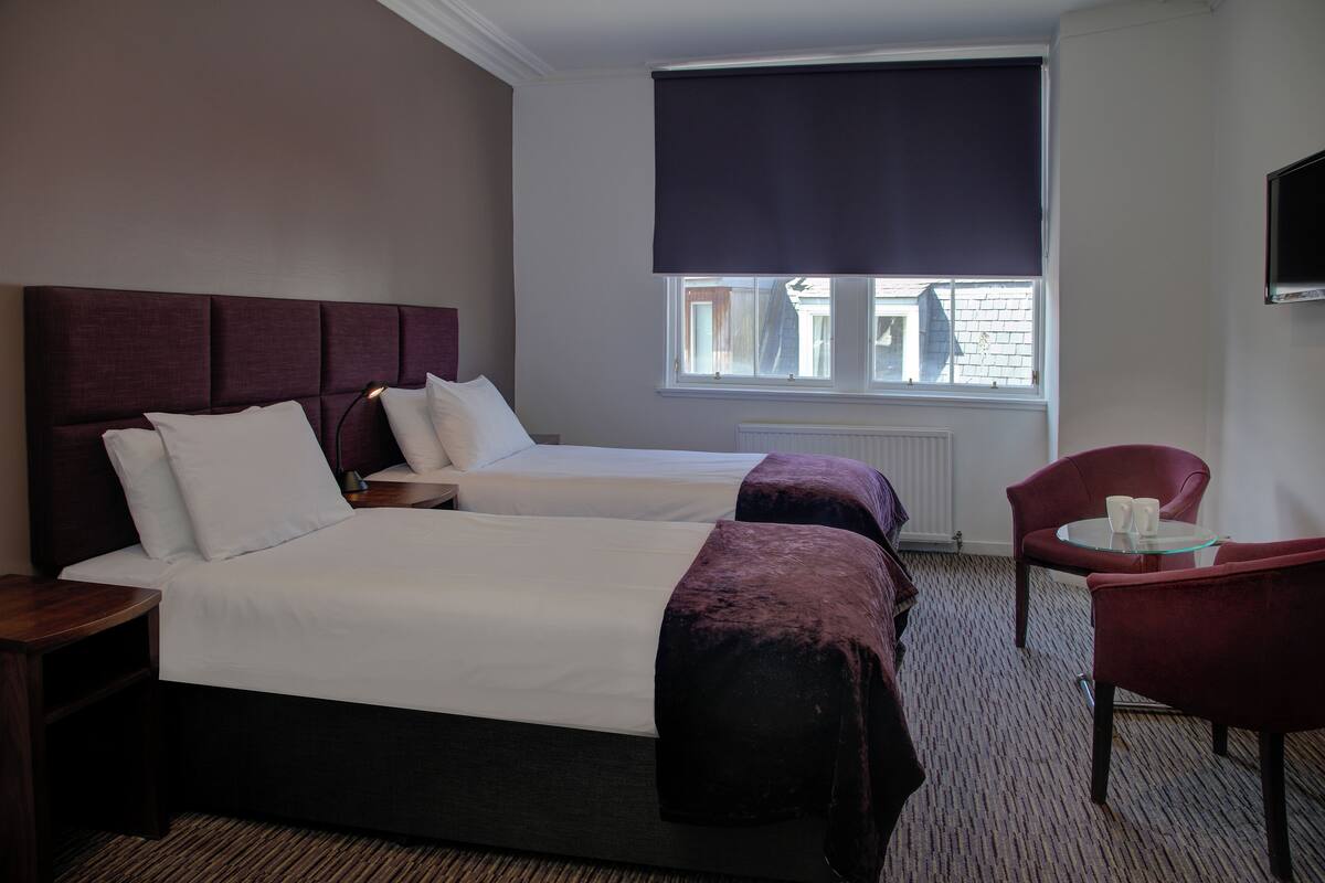 A hotel room with two beds, a sitting area with a chair and table, and a window with a dark blind.