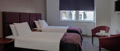 A hotel room with two beds, a sitting area with a chair and table, and a window with a dark blind.