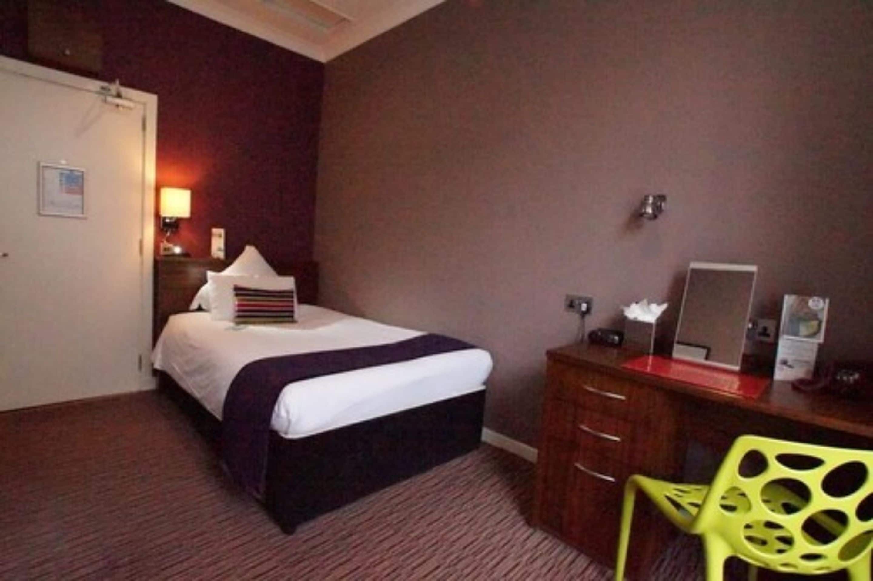 Habitación de hotel con cama, mesita de noche, lámpara, espejo y escritorio con silla.