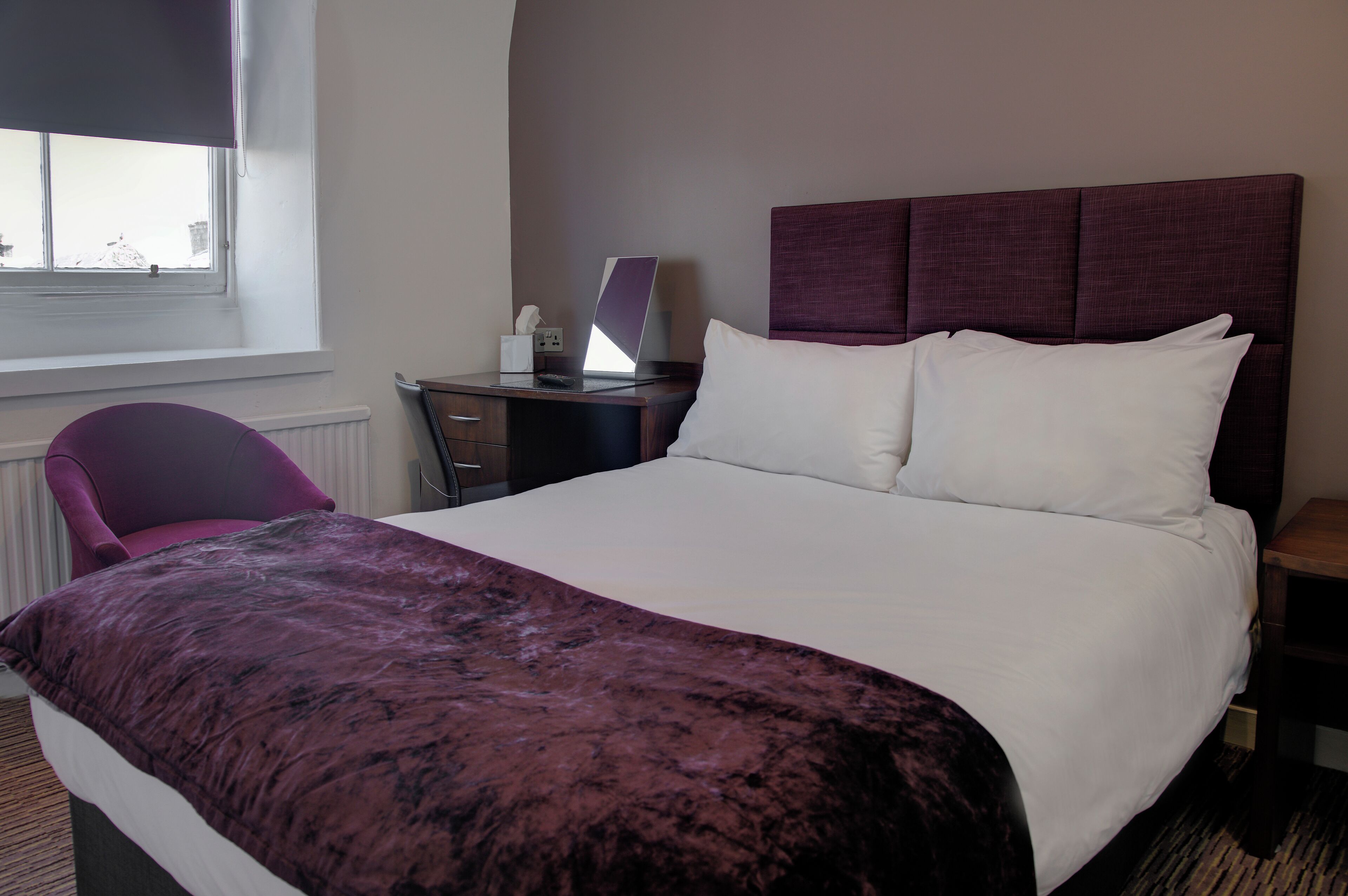 A neatly made bed with white linens and a purple headboard, a small desk with a lamp, and a window with blinds.