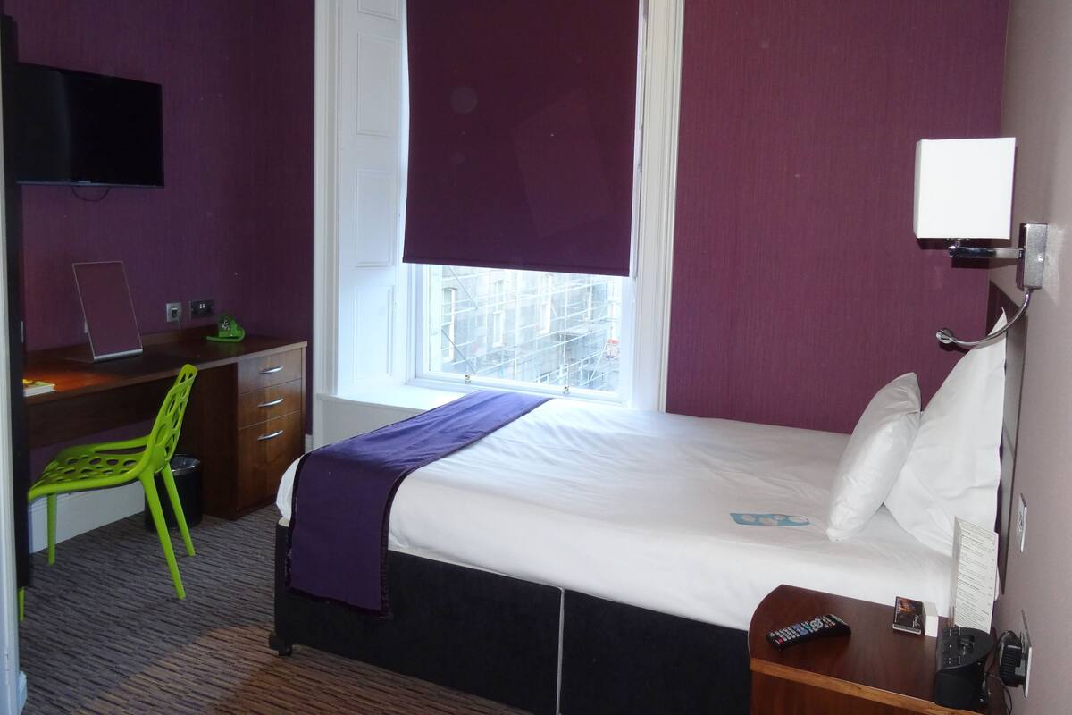 A hotel room with a bed, a desk with a chair, a TV, and a window with a view of buildings.