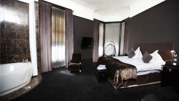 A hotel room with a large bed, a sitting chair, a bedside table, a TV, and a bathroom with a bathtub.
