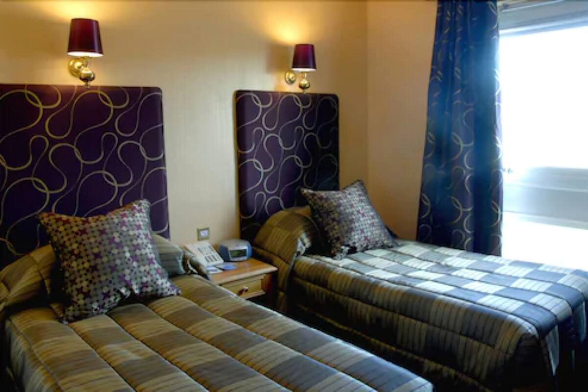 A hotel room with two beds, each with patterned headboards and matching pillows. There are bedside lamps and a telephone on the nightstand.
