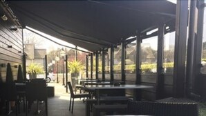 An outdoor patio area with tables and chairs, covered by a roof, and large windows offering views of the surrounding area.