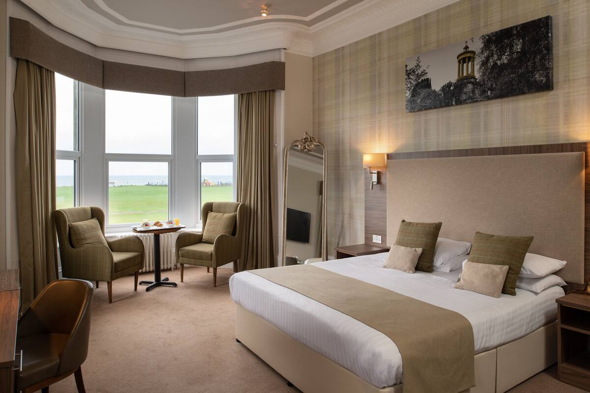 A hotel room with a large bed, two armchairs, a small table with a tray of breakfast items, a large window with curtains, and a painting on the wall.
