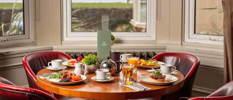 Una mesa de desayuno dispuesta con platos de comida, tazas y una tetera, con vistas al exterior a través de amplias ventanas.