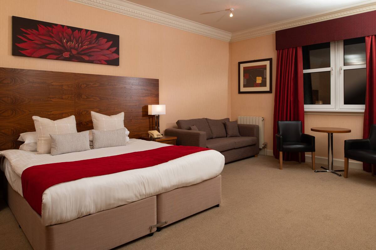 A hotel room with a large bed, a sofa, two armchairs, a small table, and a window with curtains.