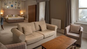 A modern living room with a beige sofa, a wooden coffee table, and a plaid armchair. There is a bed with a headboard in the background and a large window with curtains. - Fairfield House - Ayr (Ayr)