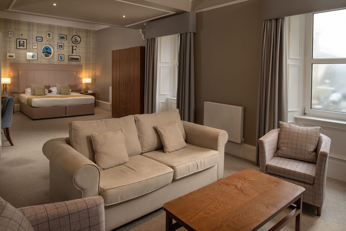 A modern living room with a beige sofa, a wooden coffee table, and a plaid armchair. There is a bed with a headboard in the background and a large window with curtains.