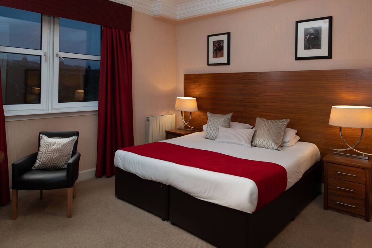 A hotel room with a large bed, two bedside tables with lamps, a chair with a cushion, and two framed pictures on the wall.