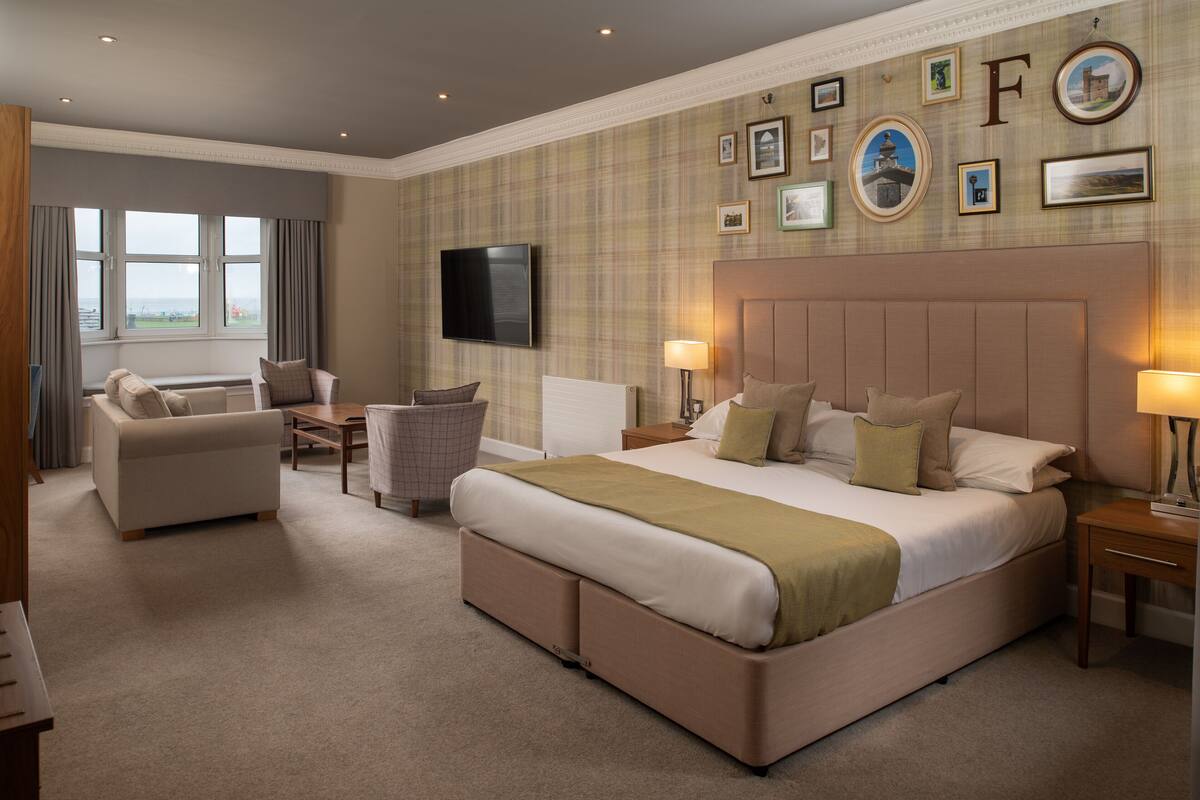 A hotel room with a large bed, a sofa, a chair, a TV, and a wall with framed pictures.