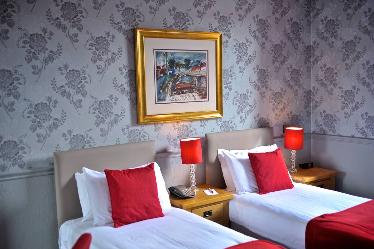 A hotel room with two beds, red and white bedding, a framed picture on the wall, and a nightstand with a lamp and phone.