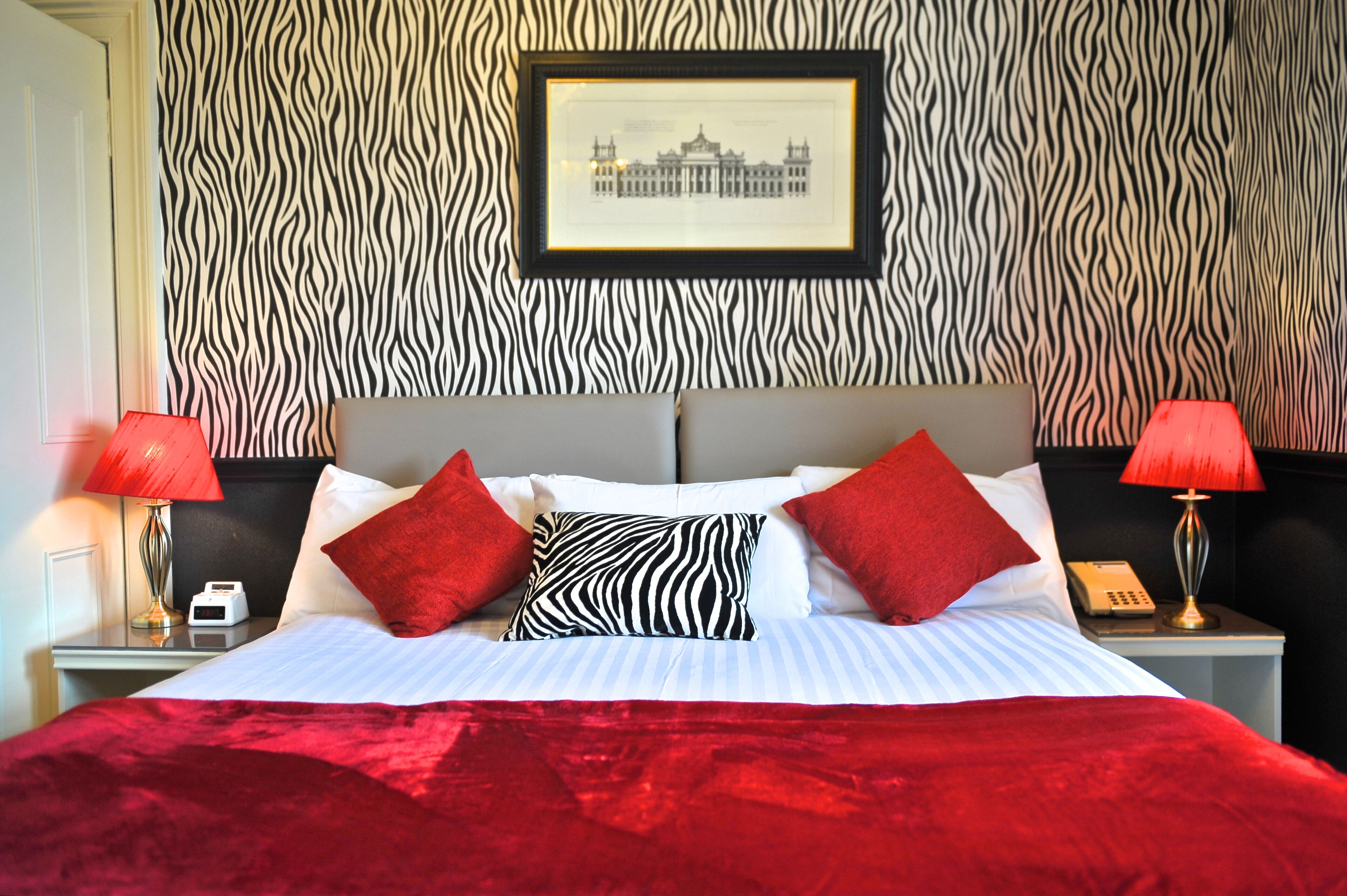 A hotel room with a zebra-patterned wall, a framed picture, a bed with red and white bedding, and two bedside lamps.
