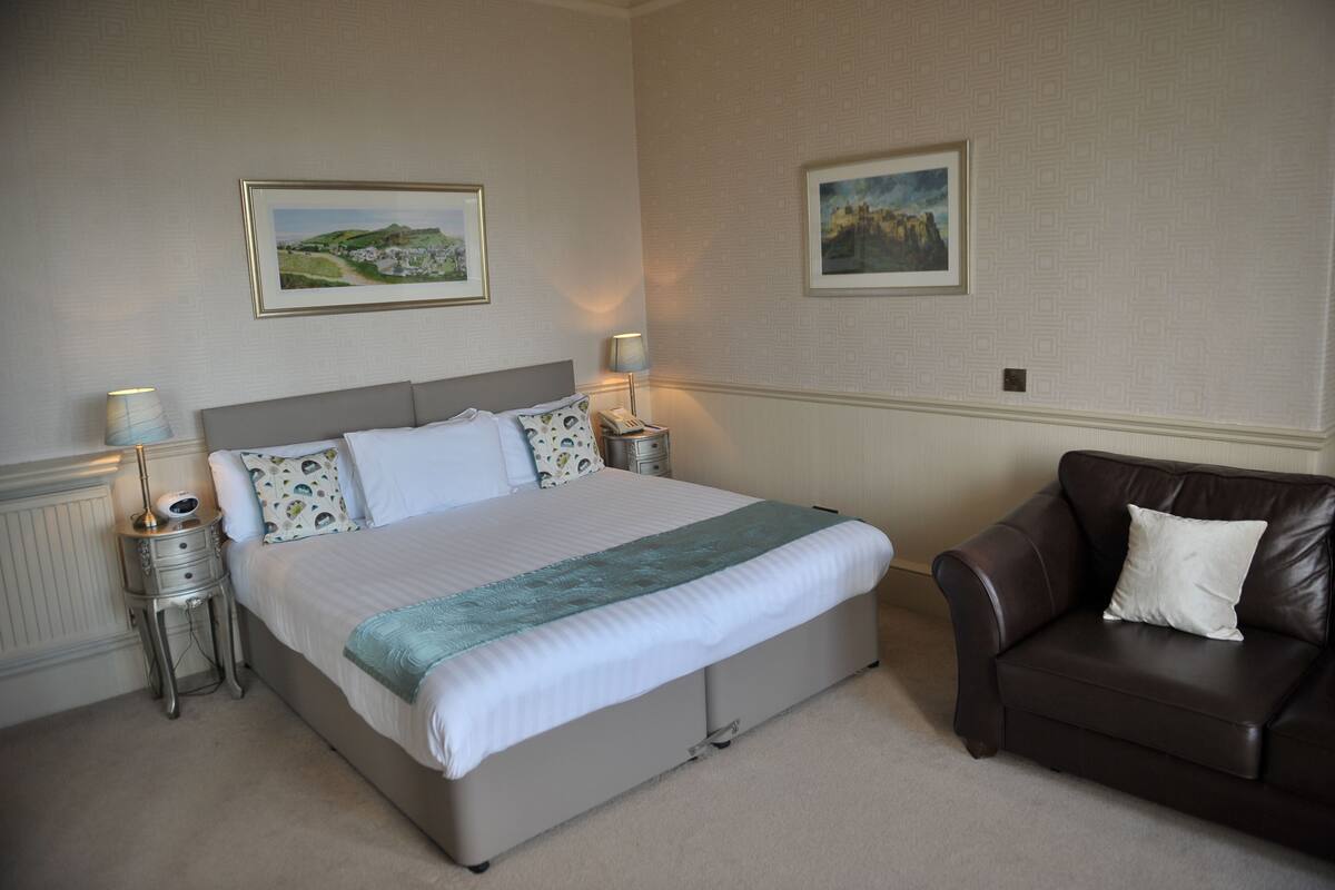 A neatly arranged bedroom with a large bed, two bedside tables, a brown leather armchair, and two framed pictures on the wall.