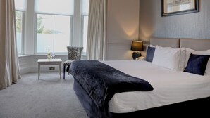 Una habitación de hotel con una cama grande, una mesita con una copa de vino, una silla y un cuadro en la pared.