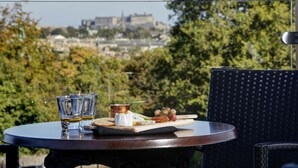 Una mesa con una tabla de quesos, copas y una botella, con vista a un paisaje urbano.