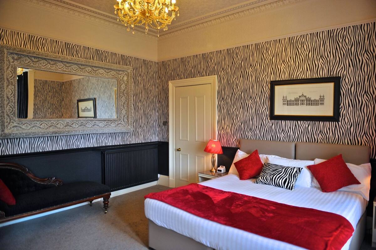 A bedroom with a zebra-striped wallpaper, a large mirror, a bed with red and black pillows, and a chandelier.