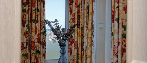 A corner room with a window, floral curtains, a vase with flowers, a wooden chair, and a small round table.