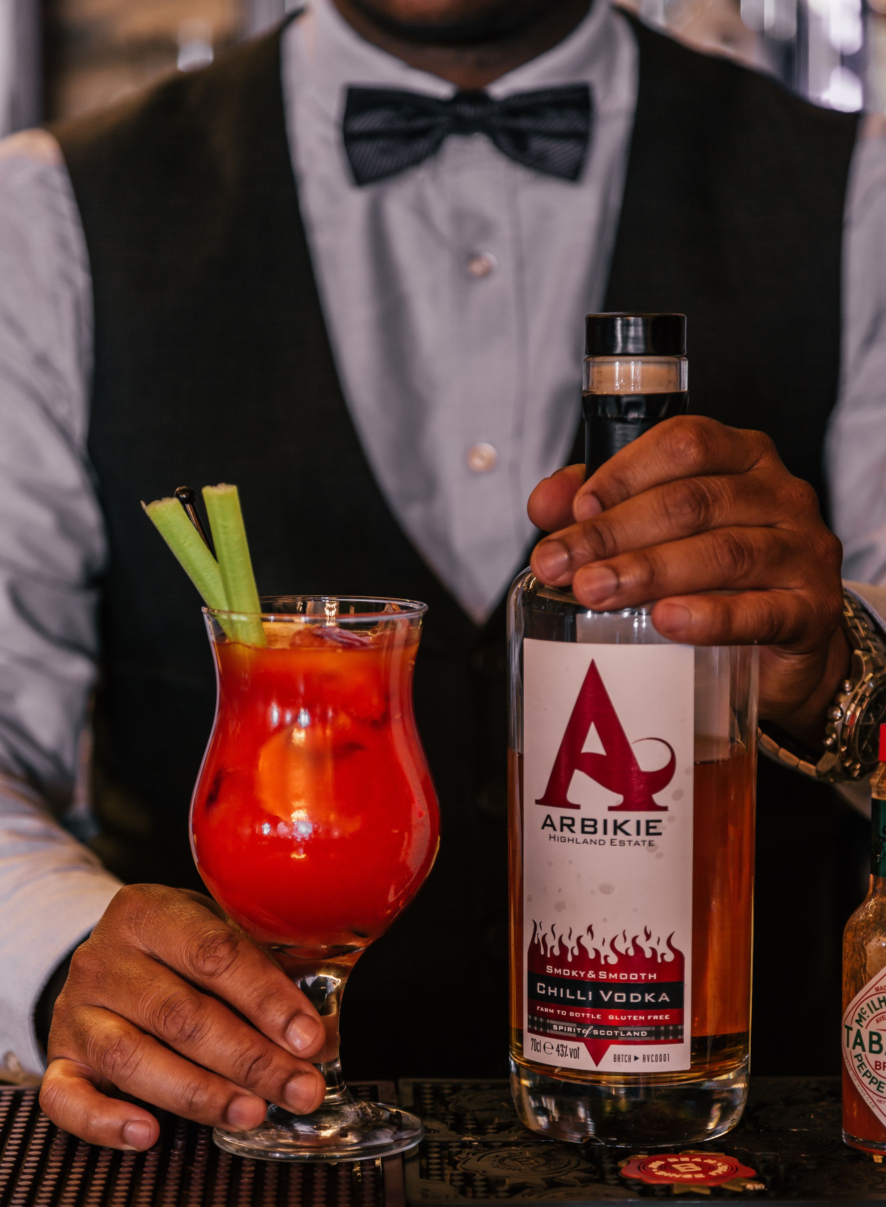 Un cantinero sosteniendo una copa de cóctel con una bebida roja y palitos de apio, y una botella de vodka Arbikie Chilli.