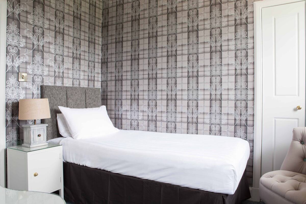A neatly made bed with white linens, a bedside lamp, and a nightstand with a lamp. The room features a plaid wallpaper pattern and a door to the right.