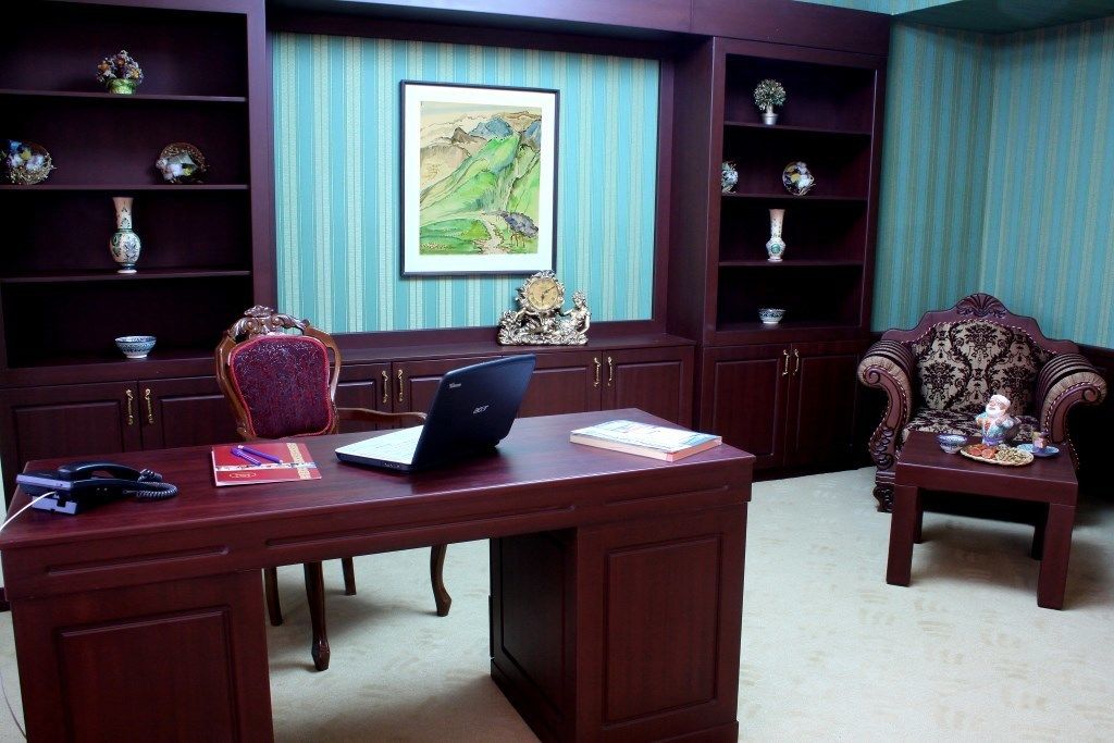 A room with a wooden desk, a laptop, and a chair. There are shelves with decorative items and a framed painting on the wall.