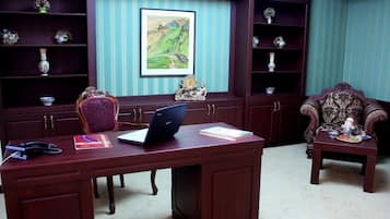 A room with a wooden desk, a laptop, and a chair. There are shelves with decorative items and a framed painting on the wall.