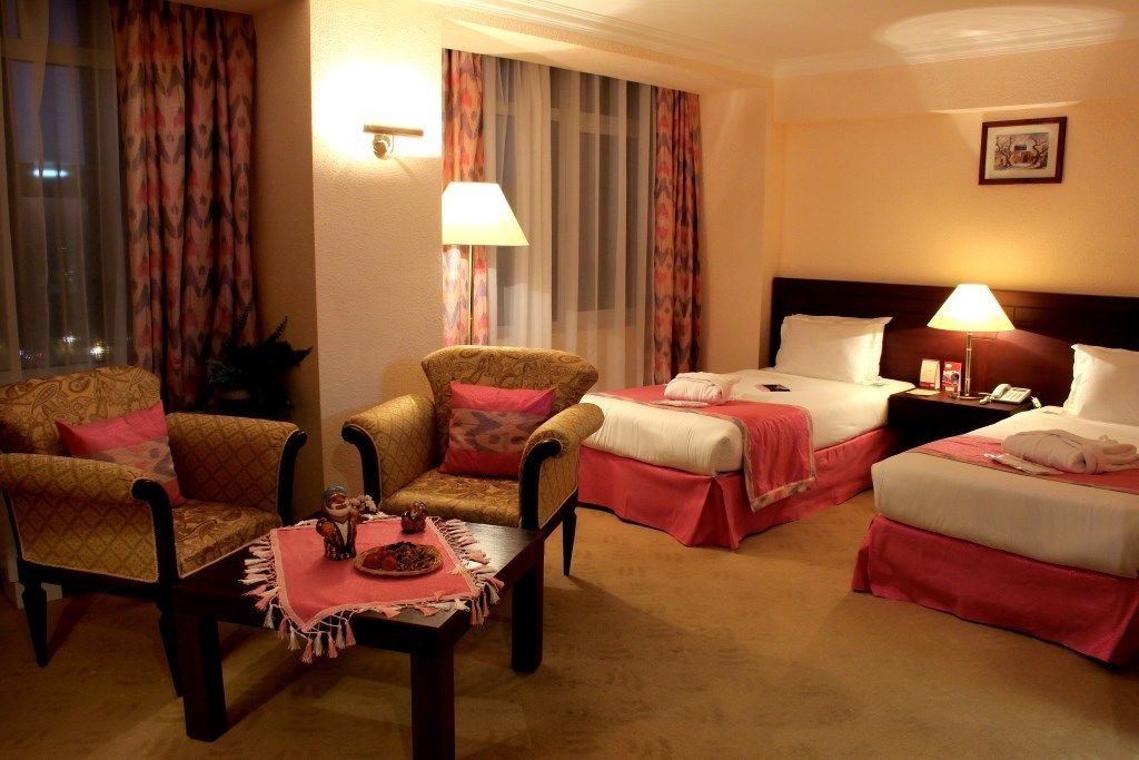 Habitación de hotel con dos camas, una zona de estar con sillas, una mesa con un centro de mesa decorativo y una mesita de noche con una lámpara.
