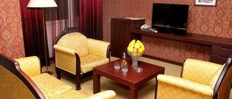 A hotel room with a sofa set, a coffee table, a TV, and a desk with a lamp.