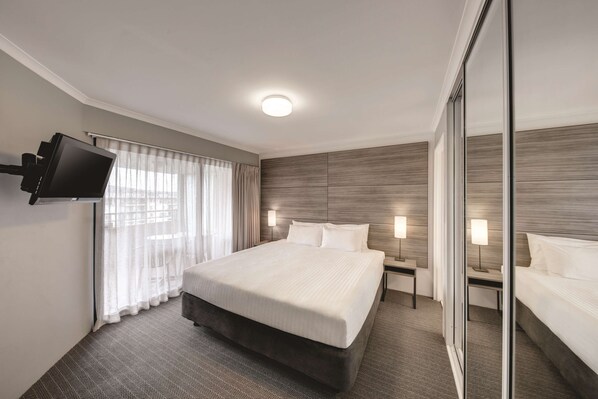 Habitación de hotel con una cama grande, un televisor de pantalla plana montado en la pared, una mesita de noche con lámpara y un espejo amplio.