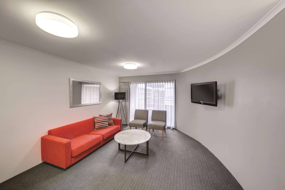 A modern living room with a red sofa, two armchairs, a coffee table, a flat-screen TV, and large windows with sheer curtains.