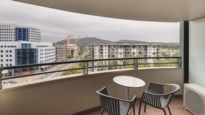 A balcony with a table and two chairs, overlooking a cityscape with modern buildings and a distant mountain.