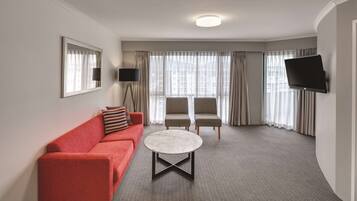 Una habitación de hotel moderna con un sofá rojo, una mesa de centro redonda, dos sillones, un televisor de pantalla plana montado en la pared y un ventanal con cortinas.