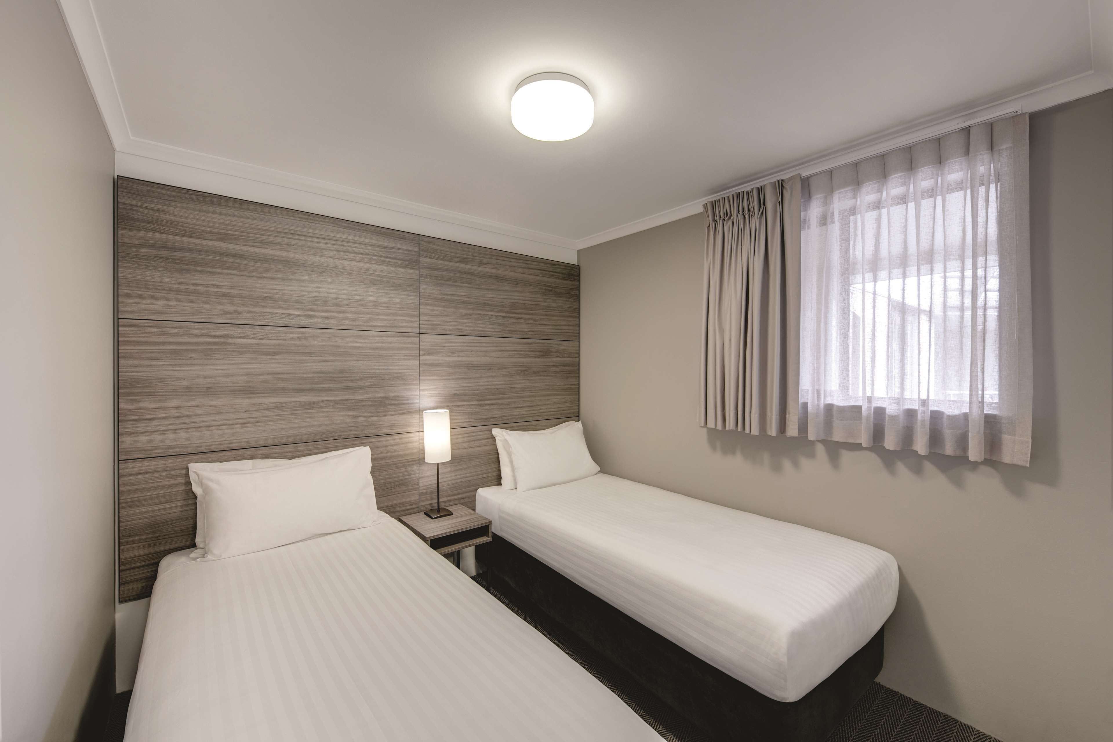A hotel room with two single beds, a nightstand with a lamp, a wooden headboard, and a window with sheer curtains.