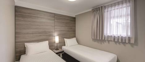 A hotel room with two single beds, a nightstand with a lamp, a wooden headboard, and a window with sheer curtains.