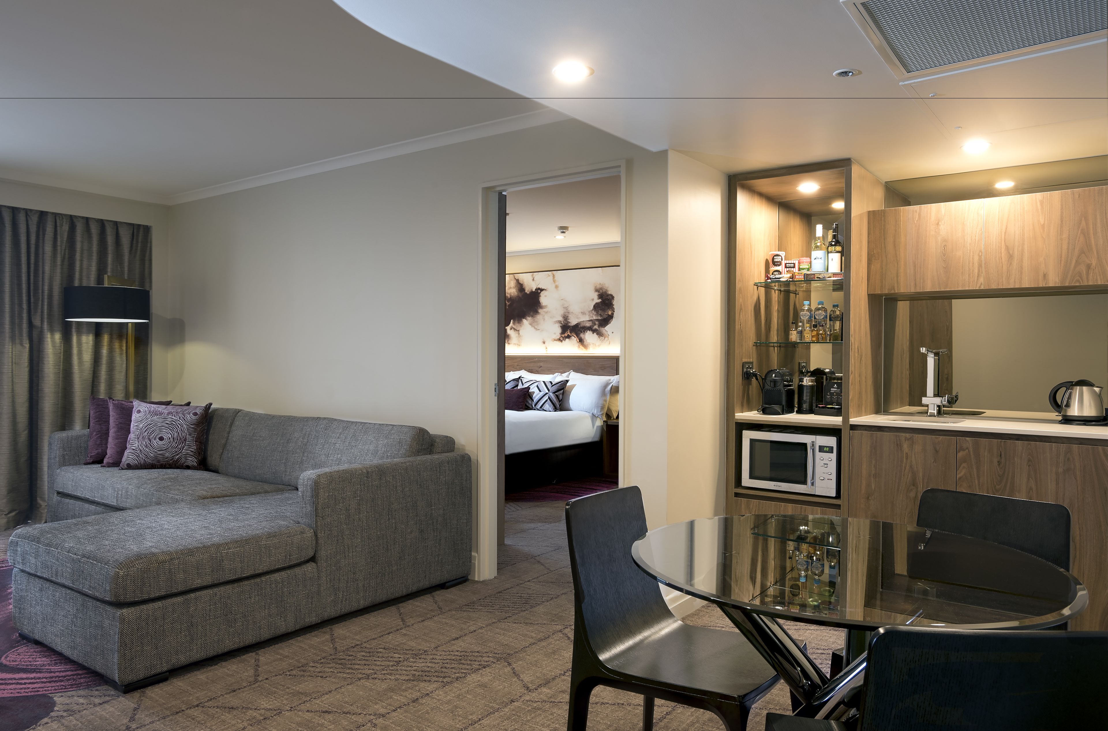 A modern hotel room with a grey sofa, a glass dining table, and a kitchenette area.
