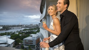 A couple standing by a window with a city view, holding champagne glasses. - Rydges South Bank (South Brisbane)