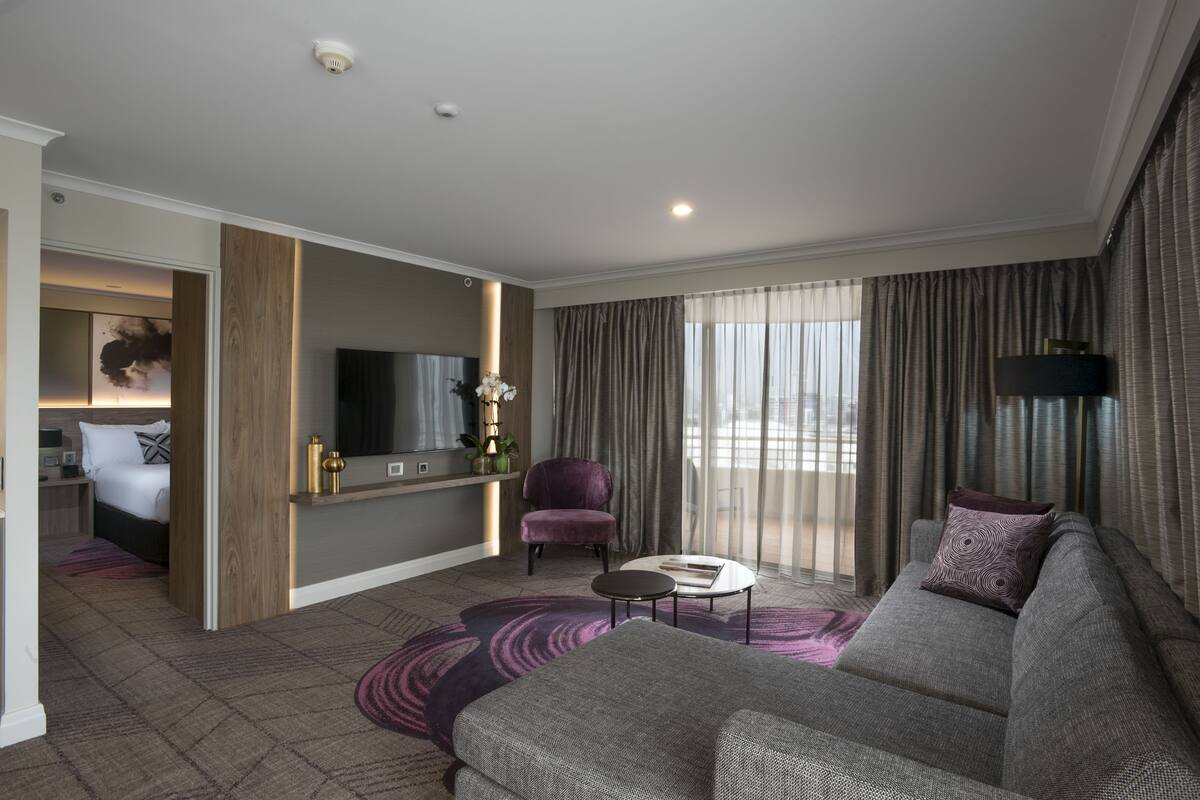 A modern hotel room with a grey sofa, a small round table, a purple armchair, a flat-screen TV, and a large window with curtains.