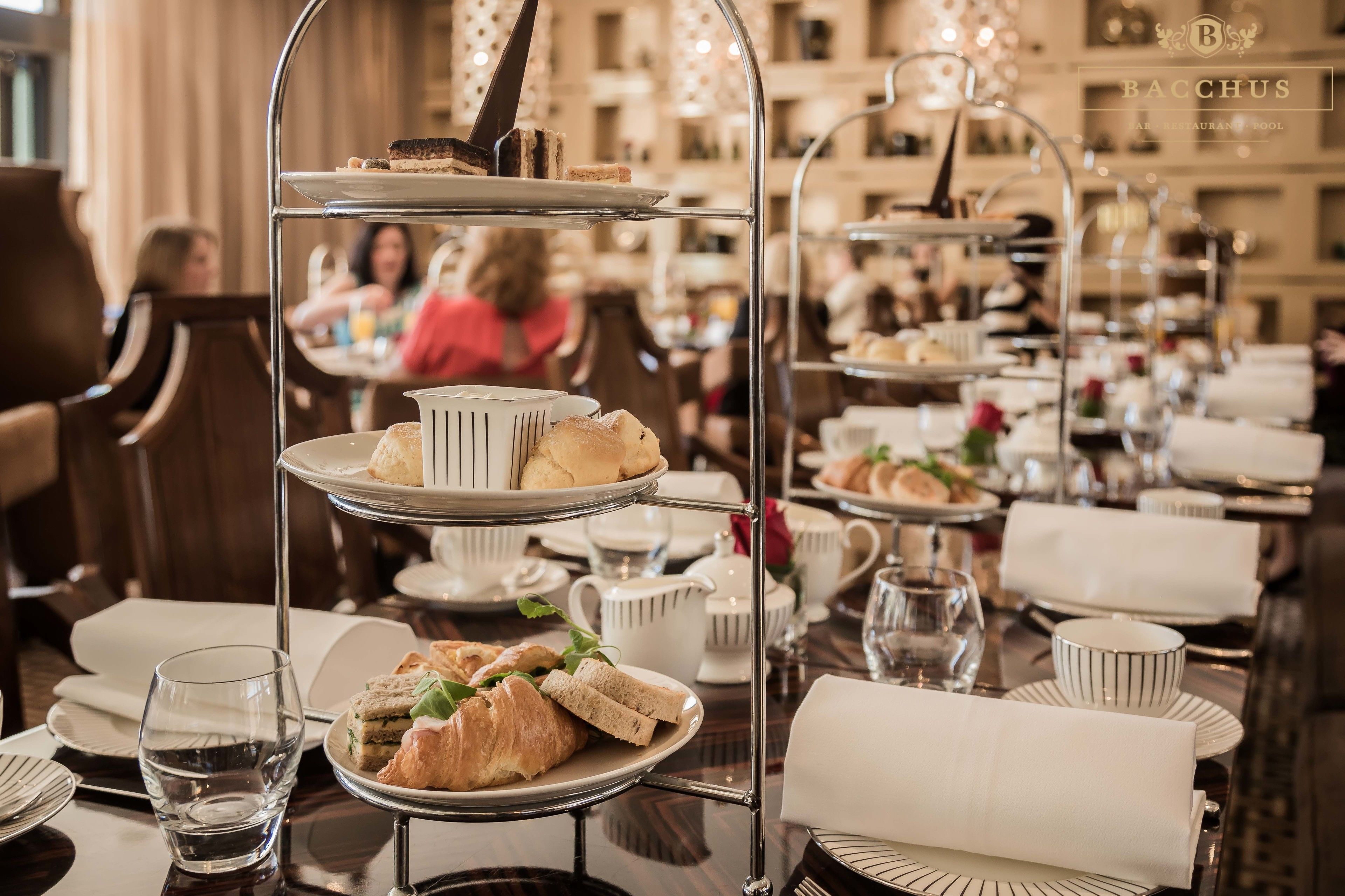 Un soporte escalonado para postres con pasteles y sándwiches, rodeado de mesas de comedor con manteles blancos y cubiertos.