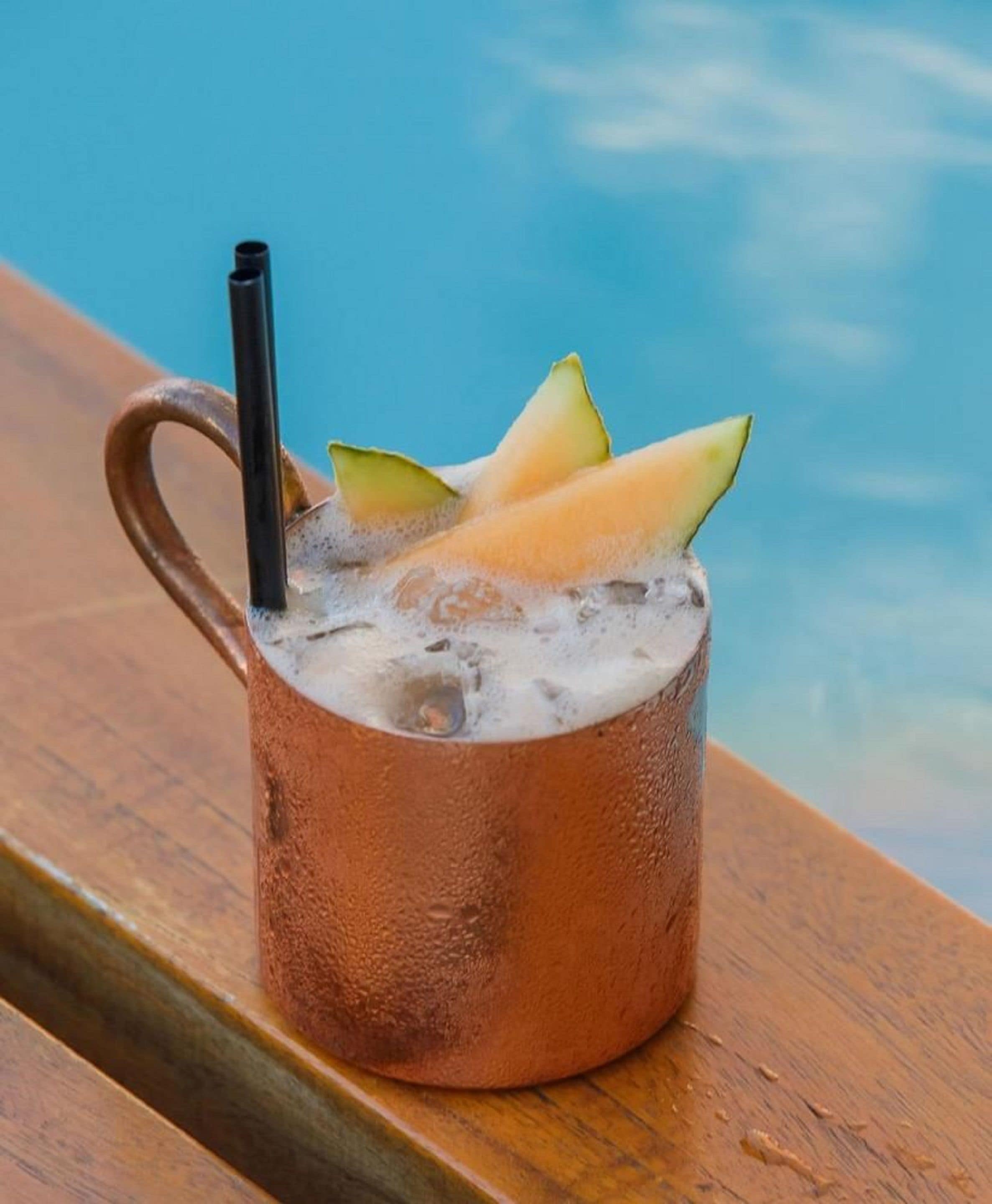 A copper mug with a handle, filled with a beverage and ice cubes, placed on a wooden surface with a blue background.