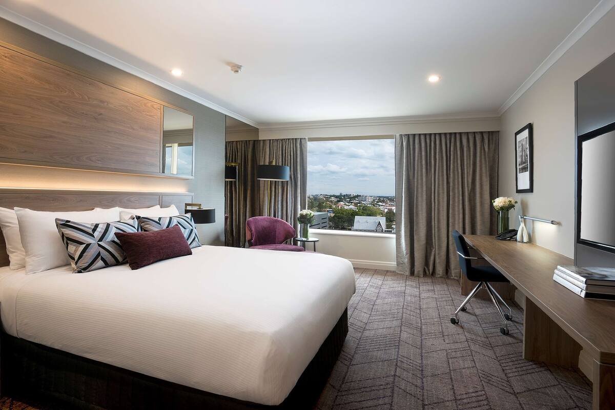 A hotel room with a large bed, a desk, a chair, a TV, and a view of the city.
